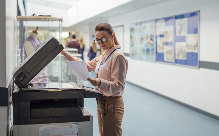 Female teacher standing next to printer in school hallway lifting up the top part of the machine looking at a paper.