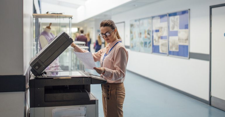 Female teacher standing next to printer in school hallway lifting up the top part of the machine looking at a paper.