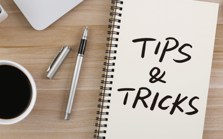Notebook on wooden desk that says tips and tricks, along with coffee cup, pens, a plant, and a smart phone