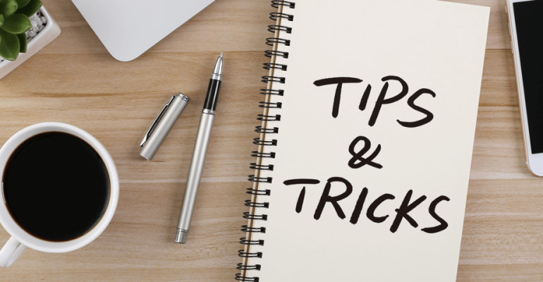 Notebook on wooden desk that says tips and tricks, along with coffee cup, pens, a plant, and a smart phone