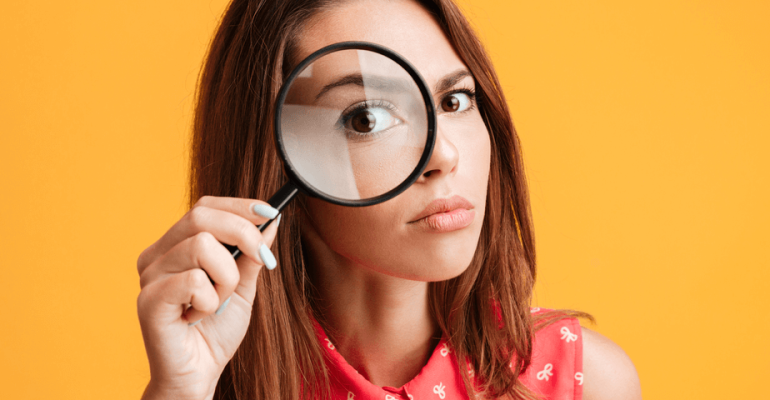 Close-up portrait of young serious woman looking through a magnifying glass, isolated over yellow background