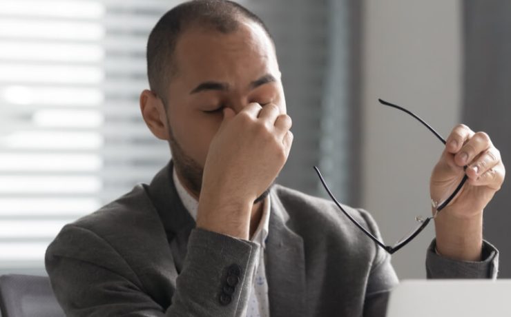 Businessman seated at desk holding his glasses in one hand with his eyes closed and the other hand holding his nose