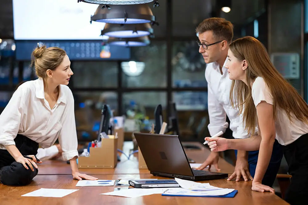 Three people around a desk in an office leaning on it and talking