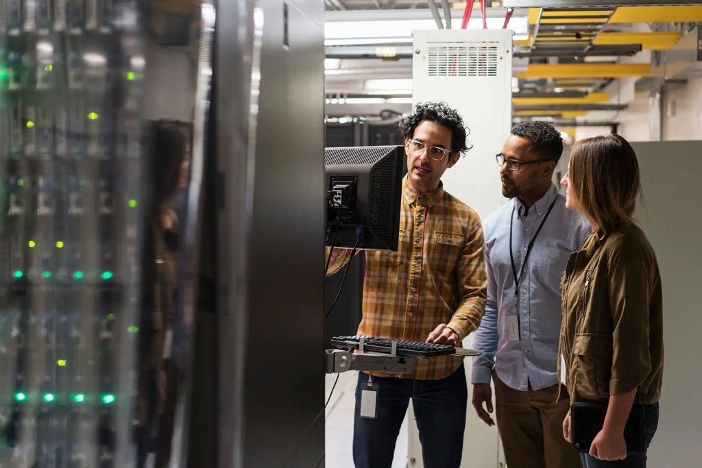 Three data center technicians standing in a server room one is working on a keyboard and they are all looking at a monitor