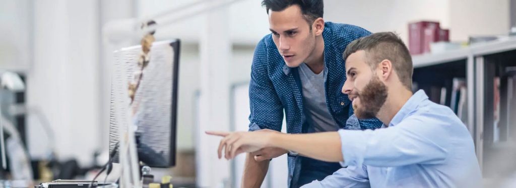 Two people at a desk one is pointing a finger at a computer monitor