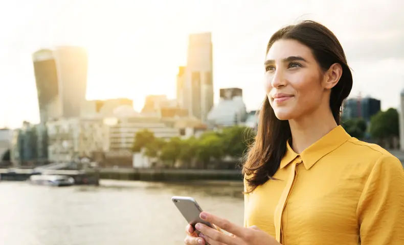 Woman outdoors holding a phone and smiling by a river there is a city skyline in the background