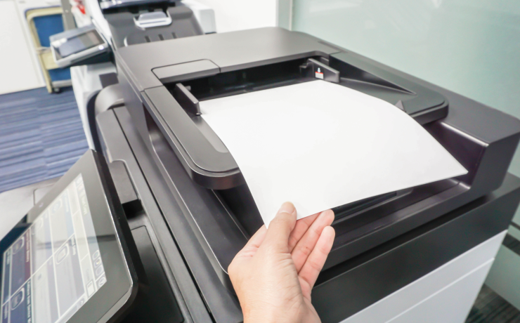 A hand feeds a paper document into the scanner of a modern office multifunction printer with a touchscreen control panel