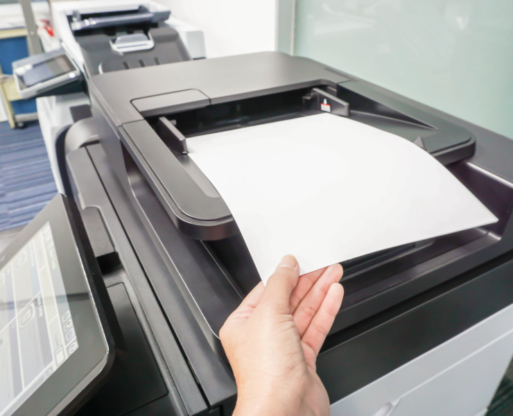 A hand feeds a paper document into the scanner of a modern office multifunction printer with a touchscreen control panel